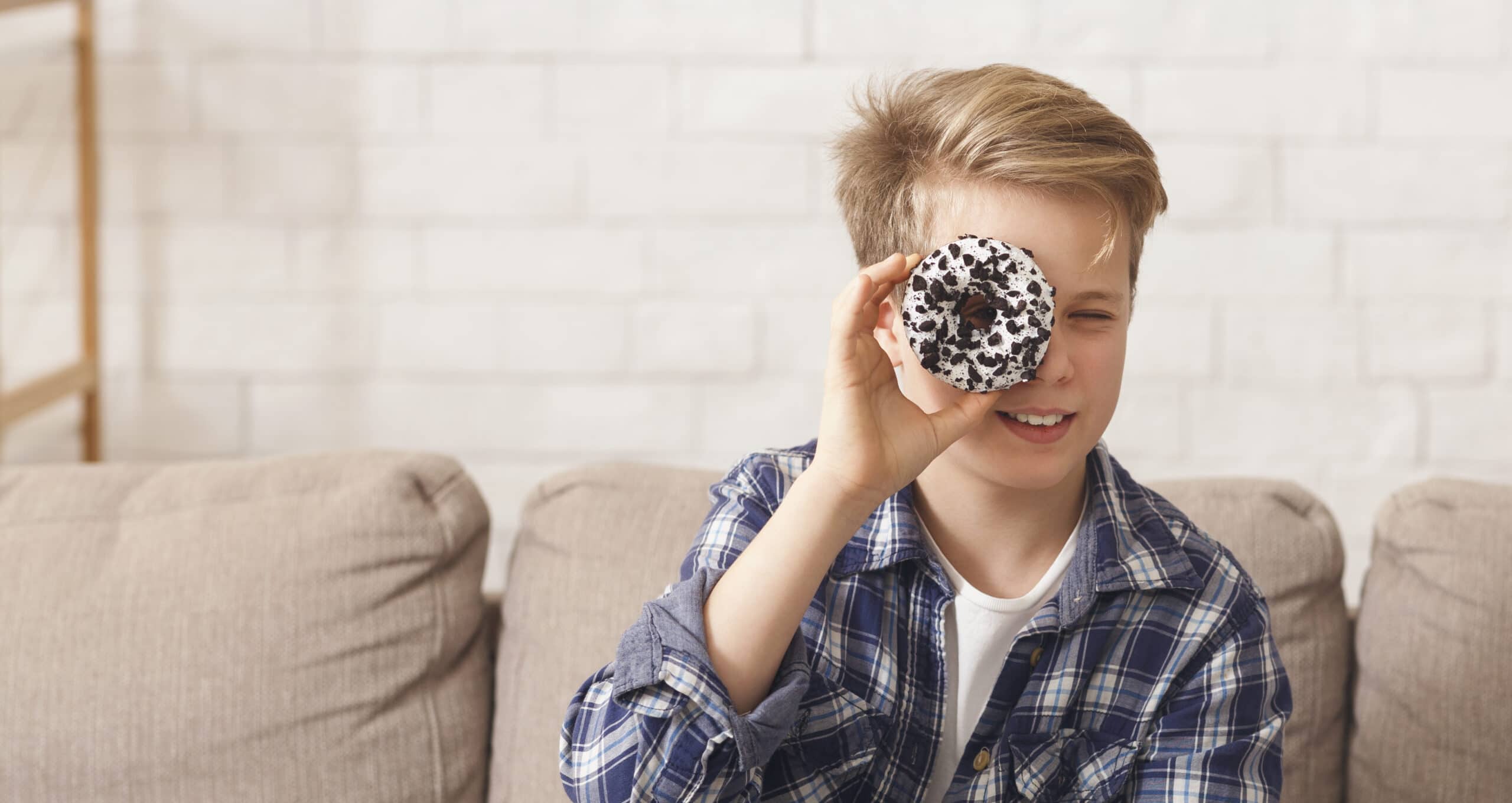Boy Holding Doughnut Near Eye Posing Sitting On Couch, Panorama